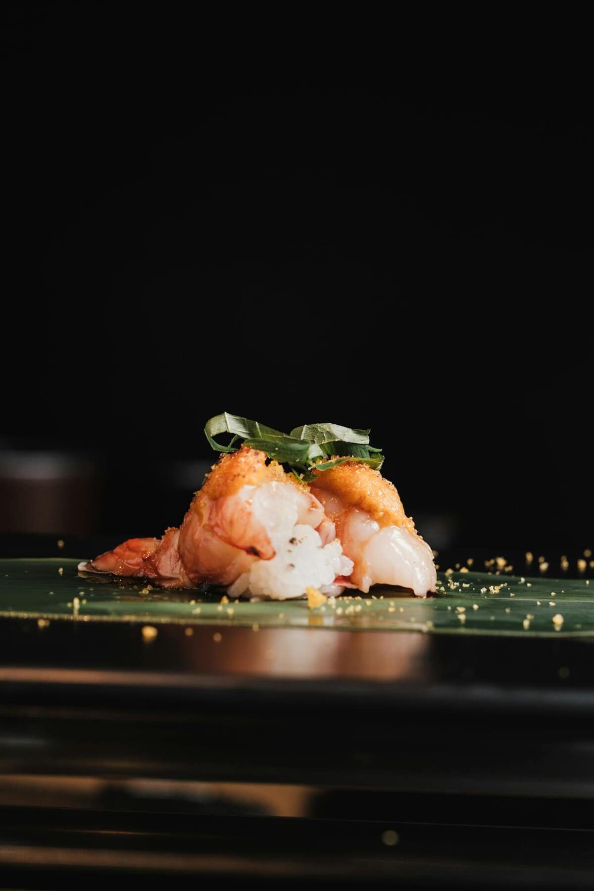 Close-up of gourmet shrimp nigiri sushi on a leaf with garnish, captured in soft lighting.