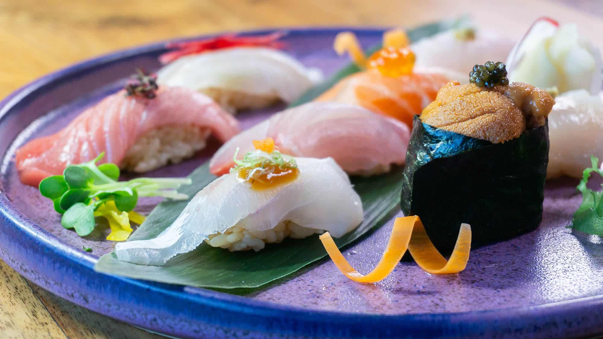 Plato de sushi Omakase con varias piezas de sushi, incluyendo nigiri y gunkan maki, servidos en un plato azul. Decorado con hierbas frescas y adornos de zanahoria. Gastronomía japonesa elegante.