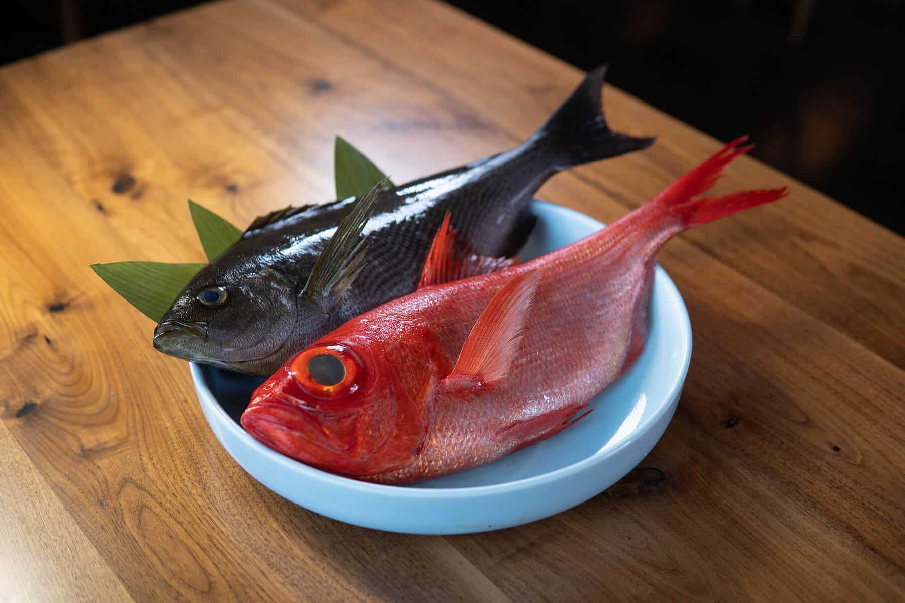 A blue bowl on a wooden table contains two whole raw fish—one silver with green-tinted fins and one bright red—each garnished with a green leaf. Both fish are displayed side by side, facing the same direction. Fuji Omakase Sushi San Fernando Valley