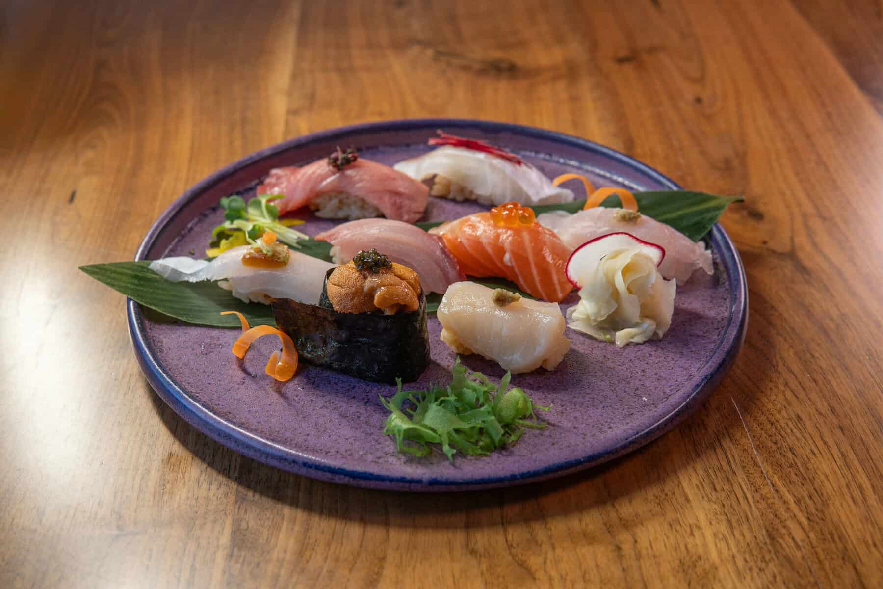 A purple plate on a wooden table holds assorted sushi pieces, including tuna, salmon, white fish, uni, and ikura. The sushi is artfully arranged on green leaves with garnishes of ginger, wasabi, and sliced vegetables. Fuji Omakase Sushi San Fernando Valley