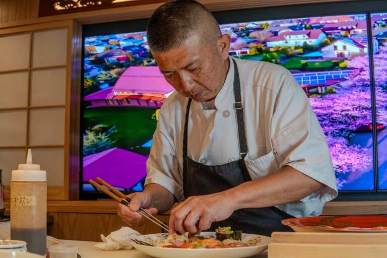 A chef in a white coat and dark apron carefully arranges sushi with chopsticks. Behind him, a large colorful screen displays a vibrant aerial view of a Japanese town with pink cherry blossoms. Bottles and dishes are on the counter in the foreground. Fuji Omakase Sushi San Fernando Valley