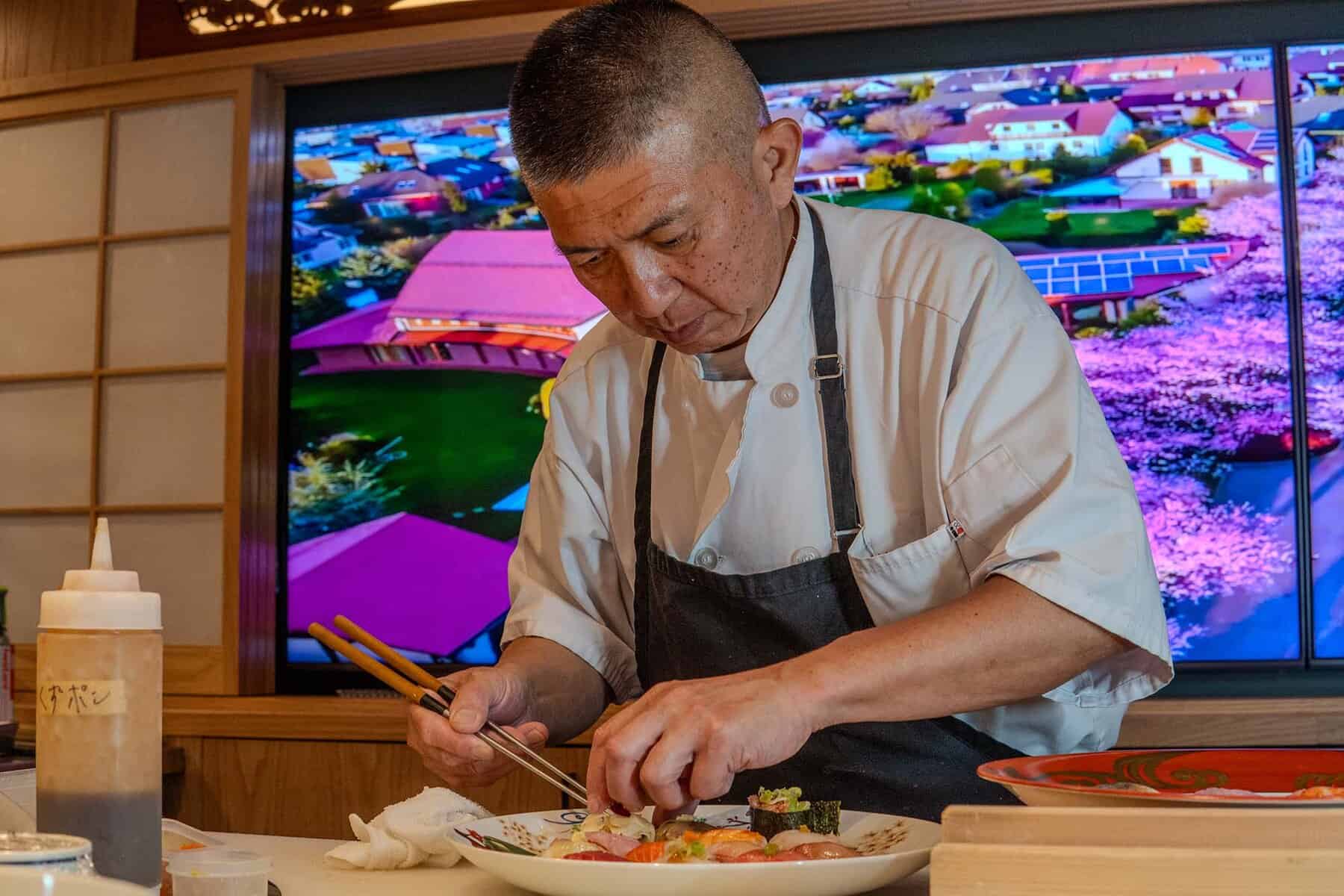 A chef in a white coat and dark apron carefully arranges sushi with chopsticks. Behind him, a large colorful screen displays a vibrant aerial view of a Japanese town with pink cherry blossoms. Bottles and dishes are on the counter in the foreground. Fuji Omakase Sushi San Fernando Valley