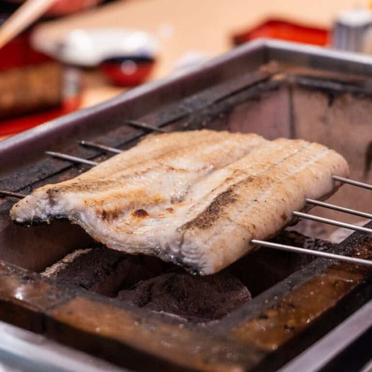 A fillet of eel is skewered with metal rods and being grilled over hot charcoal in a rectangular metal grill. The eel’s surface is slightly browned, indicating it is cooking. The background includes blurred kitchen tools and dishes. Fuji Omakase Sushi San Fernando Valley