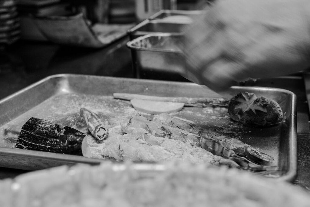 Black and white image of a metal tray holding a shrimp, a sliced vegetable, and flour. A hand reaches in from the top right, apparently preparing food. The background shows several containers and a cluttered kitchen workspace. Fuji Omakase Sushi San Fernando Valley