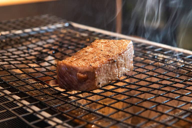 A thick, juicy steak is sizzling on a black wire grill. Wisps of steam rise, indicating it is being cooked. The steak has a seared, browned crust and is positioned at the center, with blurred background and warm lighting. Fuji Omakase Sushi San Fernando Valley