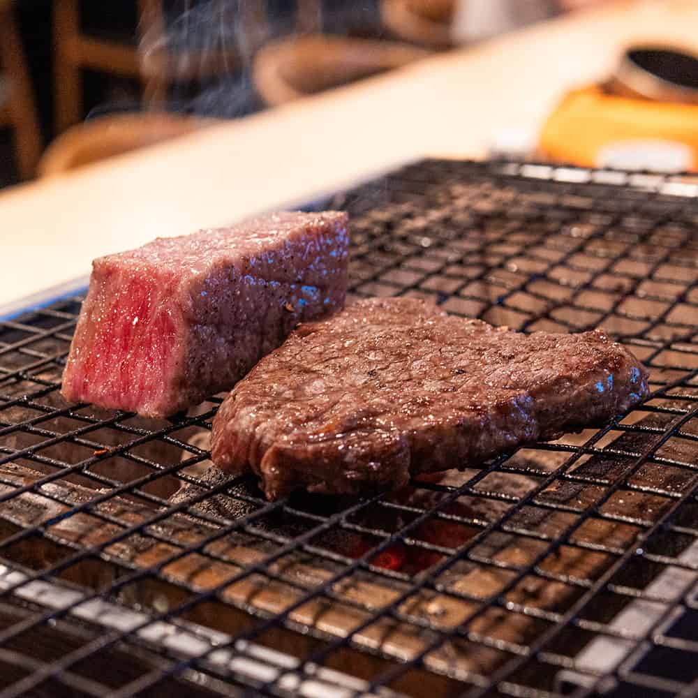 Two thick pieces of steak, one raw and marbled with fat and the other browned and cooked, sit on a wire grilling rack over an open flame in a restaurant setting. Steam rises from the hot meat, with chairs and a table blurred in the background. Fuji Omakase Sushi San Fernando Valley