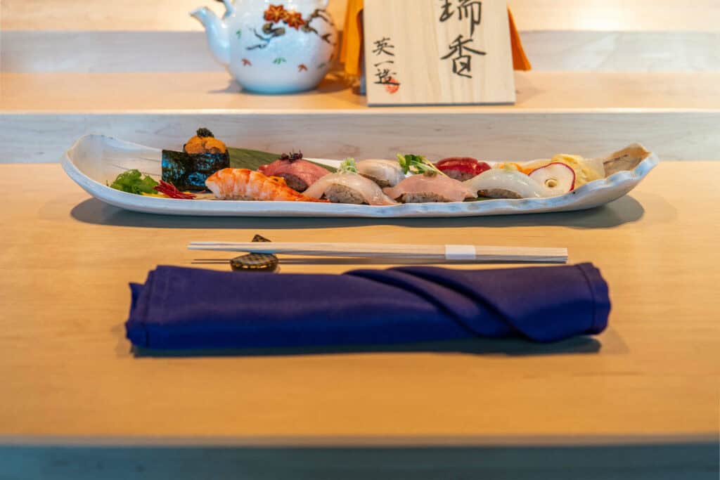 A long ceramic plate with assorted sushi pieces is set on a light wooden counter. In front, wrapped chopsticks rest on a dark blue napkin. In the background, a teapot and a wooden sign with Japanese writing evoke the spirit of Fuji Omakase. Fuji Omakase Sushi San Gabriel Valley