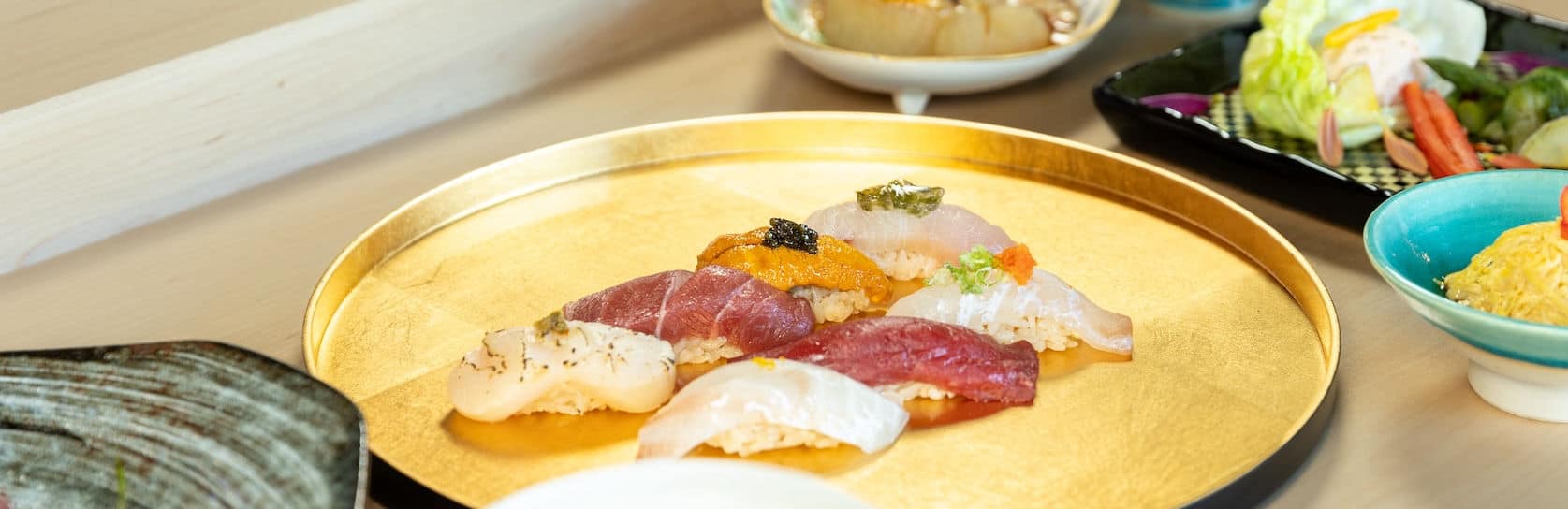 A golden tray displays assorted sushi pieces with different fish toppings, garnishes, and roe. Surrounding dishes include sliced vegetables, salad, and a bowl of light yellow noodles, all arranged on a light wooden surface. Fuji Omakase Sushi San Fernando Valley