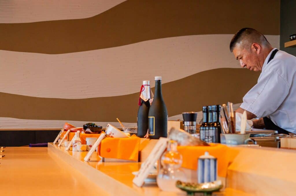 A sushi chef in a white uniform works behind a wooden counter with various dishes, bottles, and utensils. The background features a wall with wavy beige and white stripes. The atmosphere feels warm and focused. Fuji Omakase Sushi San Fernando Valley