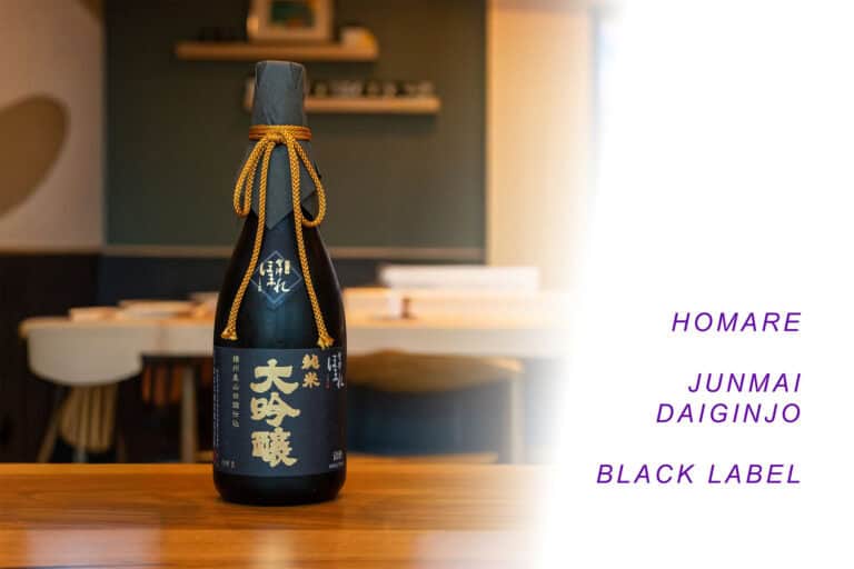 A dark bottle of Junmai Daiginjo Homare Black Labe sake with gold accents and Japanese text stands on a wooden table in a softly lit modern dining room. To the right, purple text reads: "HOMARE JUNMAI DAIGINJO BLACK LABEL" on a white background. Fuji Omakase Sushi San Fernando Valley