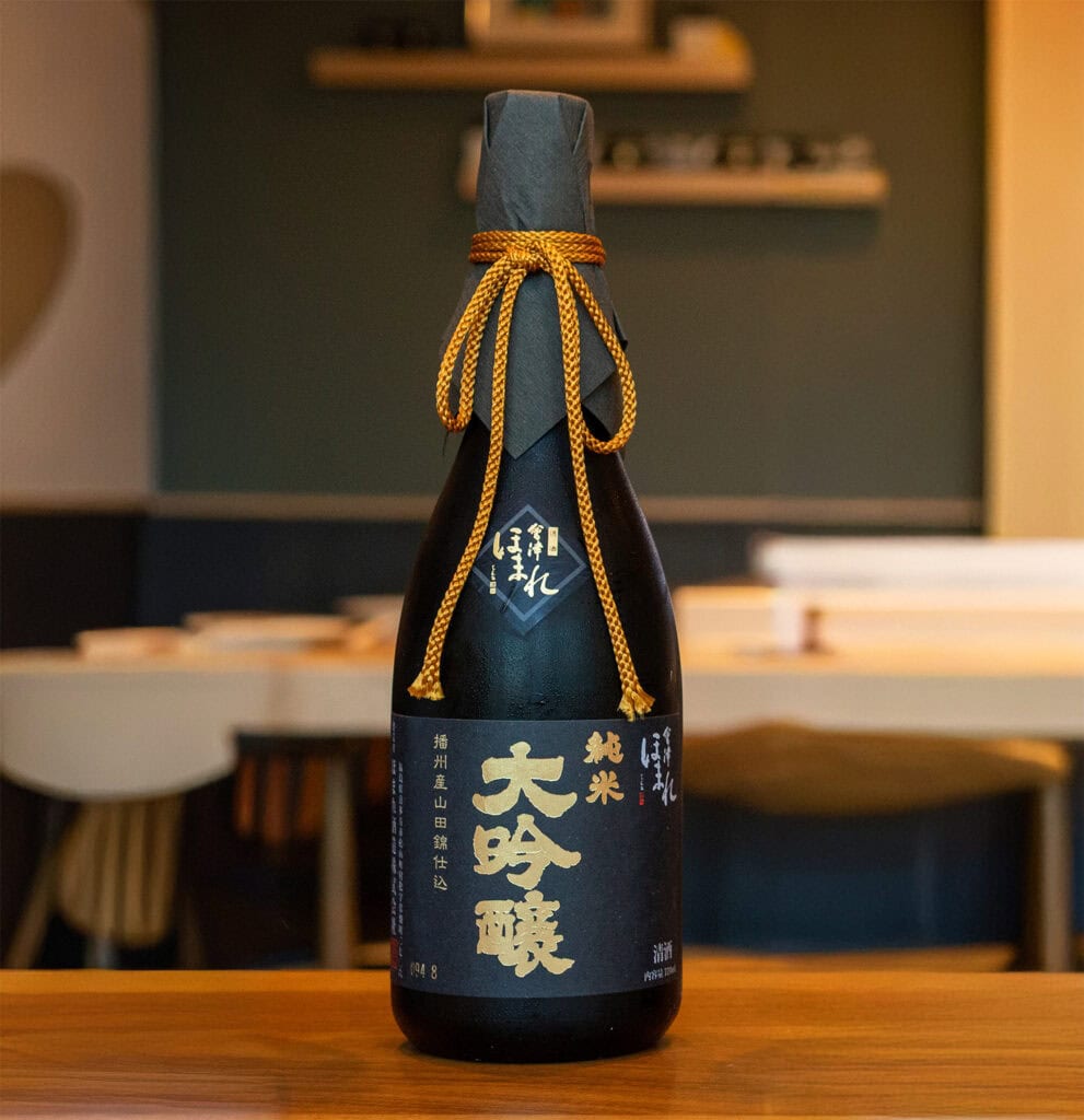 Homare Black Label Sake bottle with gold and white Japanese calligraphy on the label sits on a wooden table. The bottle is wrapped in black paper with a decorative gold cord tied around the neck. A softly lit, modern interior with shelves is in the background. Fuji Omakase Sushi San Fernando Valley