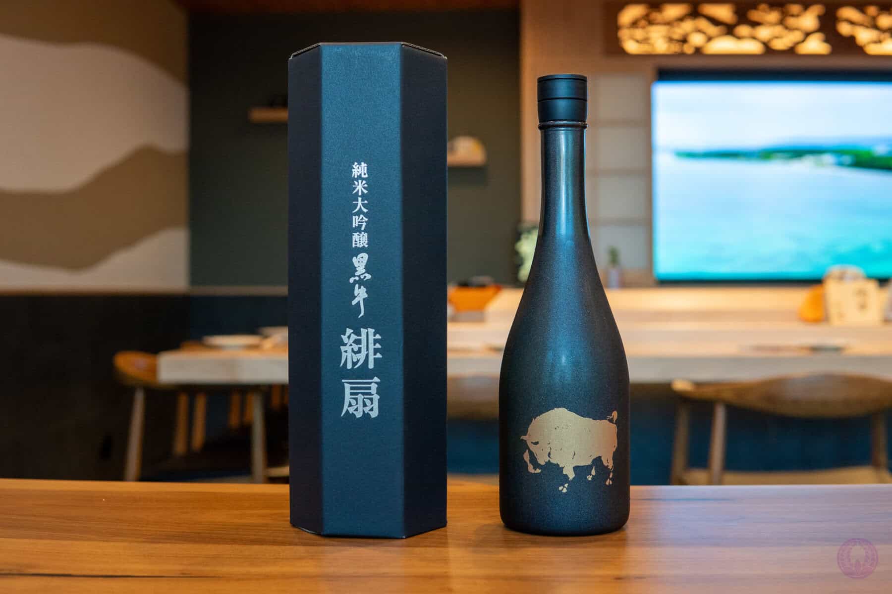 A sleek, matte black sake bottle of Kuroushi Hiougi Junmai Daiginjo with a gold map design sits on a wooden table next to a tall, matching black box with white Japanese characters. The background shows a modern, softly lit room with wooden furniture and a large screen displaying a scenic view. Fuji Omakase Sushi San Fernando Valley