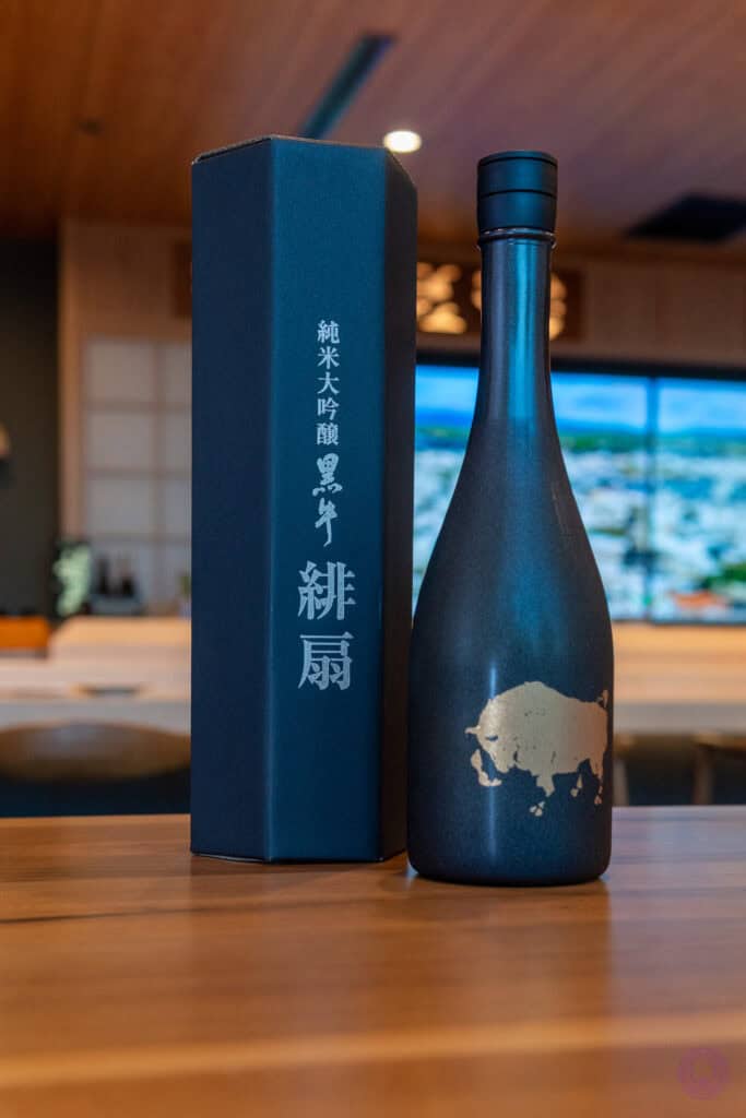 A sleek dark sake bottle with a gold buffalo design stands on a wooden table next to an upright black box featuring Japanese characters, in a modern bar with blurred seating and a digital landscape screen in the background. Fuji Omakase Sushi San Fernando Valley