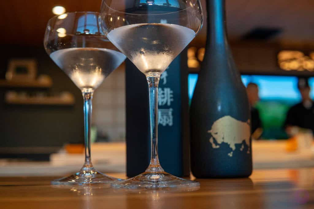 Two elegant cocktail glasses filled with a clear, chilled liquid sit on a wooden table. Behind them are two black bottles, one with Japanese characters and the other featuring a gold buffalo illustration, in a warmly lit modern room. Fuji Omakase Sushi San Fernando Valley