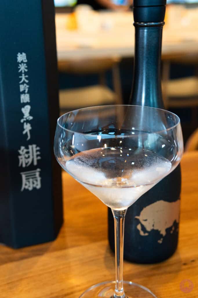 A sleek black sake bottle and its box with Japanese writing stand behind a clear, elegant cocktail glass filled with chilled sake. The glass shows condensation, resting on a polished wooden table, suggesting a refined and sophisticated drinking experience. Fuji Omakase Sushi San Fernando Valley