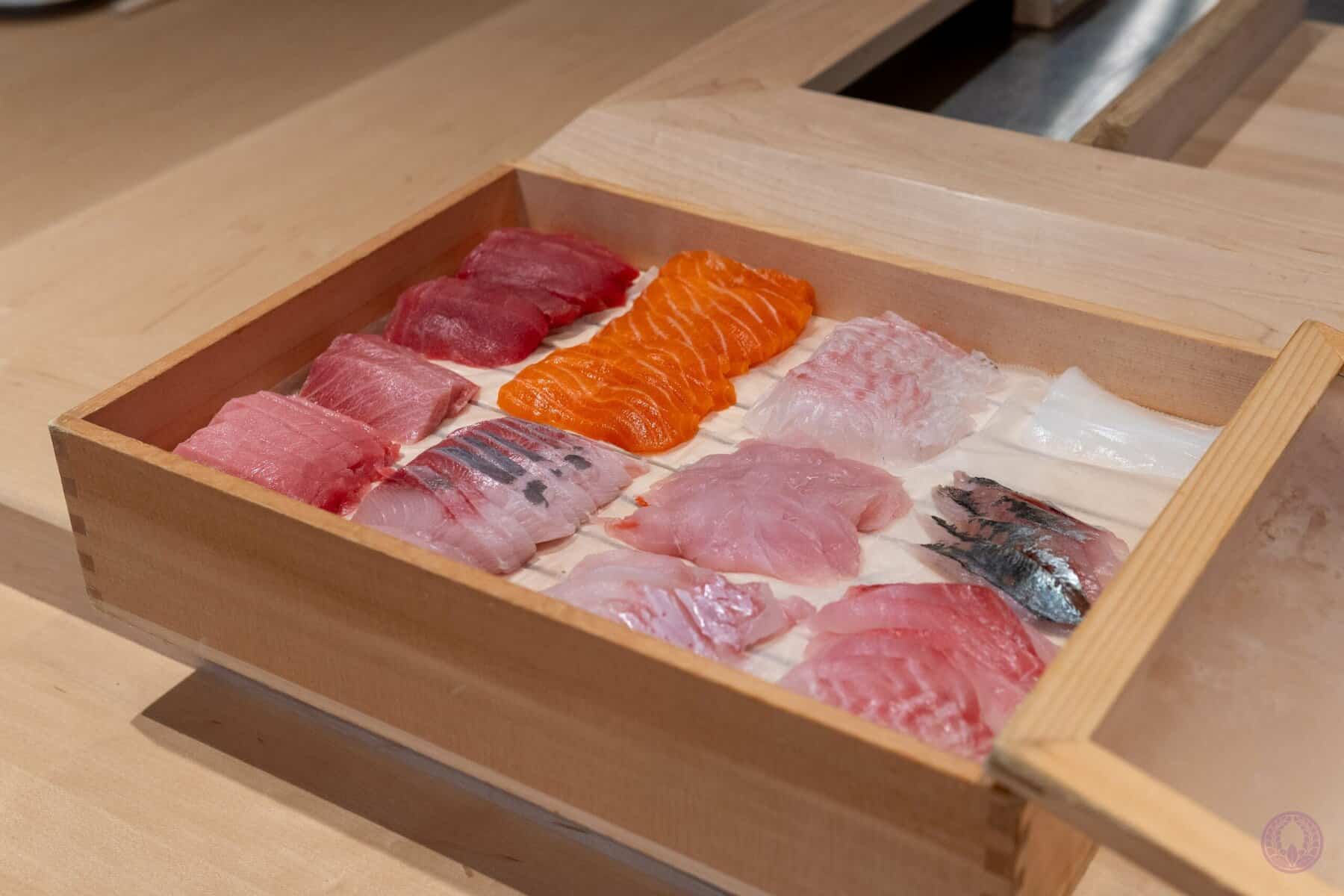 A wooden box displays an assortment of sashimi slices, including tuna, salmon, white fish, and mackerel, neatly arranged in rows on a light wooden counter, showcasing a variety of colors and textures. Fuji Omakase Sushi San Fernando Valley