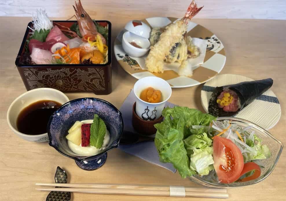 A Japanese meal set on a wooden table includes sashimi, tempura, salad with lettuce and tomato, tofu with a strawberry, dipping sauces, a hand roll with fish roe, and chawanmushi, accompanied by chopsticks and a spoon. Fuji Omakase Sushi San Fernando Valley