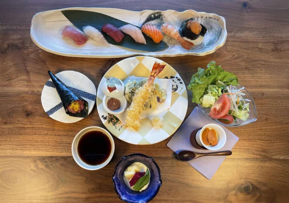 A Japanese meal set on a wooden table includes assorted nigiri sushi on a long plate, a mixed tempura dish, a salad with tomatoes and lettuce, a hand roll, dipping sauce, a small dish of uni (sea urchin), and a dessert with a green garnish. Fuji Omakase Sushi San Fernando Valley