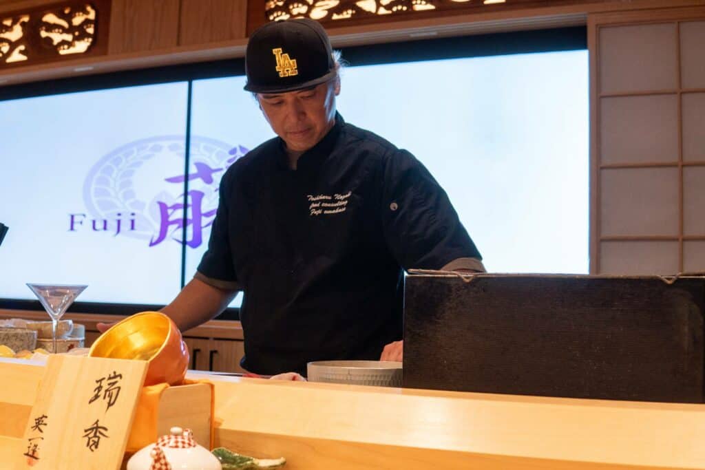 A sushi chef in a black uniform and cap prepares food behind a wooden sushi bar. Japanese decor and food items are visible, with a large screen in the background displaying the word &quot;Fuji&quot; and Japanese characters. Fuji Omakase Sushi San Fernando Valley