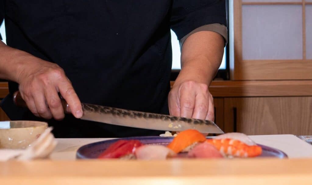 A chef in a dark uniform skillfully slices ingredients with a large knife on a white cutting board. In the foreground, assorted sushi pieces are arranged on a blue plate. Wooden panels and a shoji screen are visible in the background. Fuji Omakase Sushi San Fernando Valley