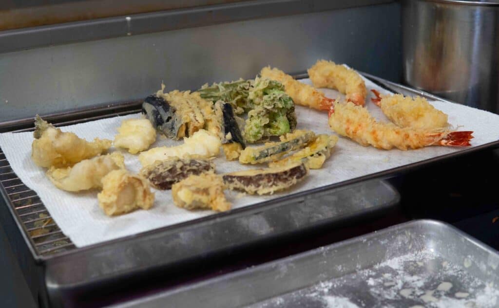 A variety of freshly fried tempura, including shrimp, fish, and assorted vegetables, is placed on a paper towel-lined wire rack to drain excess oil in a kitchen setting. A metal tray is visible below to catch any oil drips. Fuji Omakase Sushi San Fernando Valley