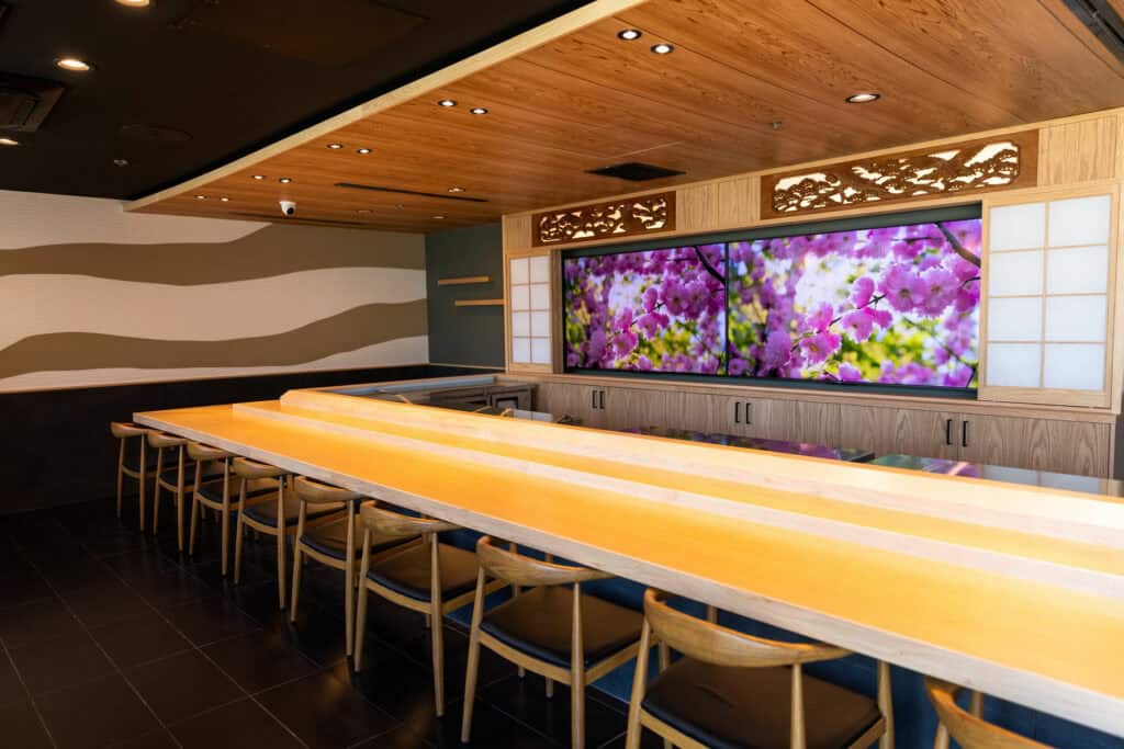 A modern Japanese restaurant with a long wooden counter, wooden chairs, and a ceiling with wood paneling. Behind the counter is a large wall screen showing cherry blossoms. Soft lighting and minimalist decor create a calm atmosphere. Fuji Omakase Sushi San Fernando Valley