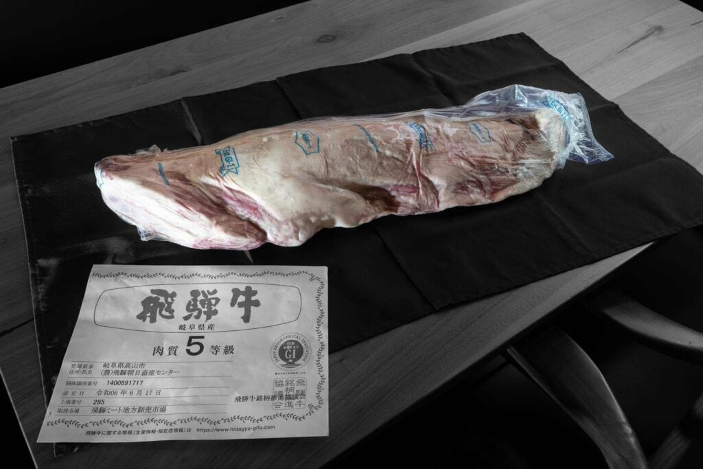 A large, raw cut of Japanese Wagyu beef in vacuum-sealed plastic rests on a black cloth atop a wooden table. Beside it, a certification paper with Japanese text and grading number &ldquo;5&rdquo; emphasizes its authenticity. The meat stands out in vivid color. Fuji Omakase Sushi San Fernando Valley