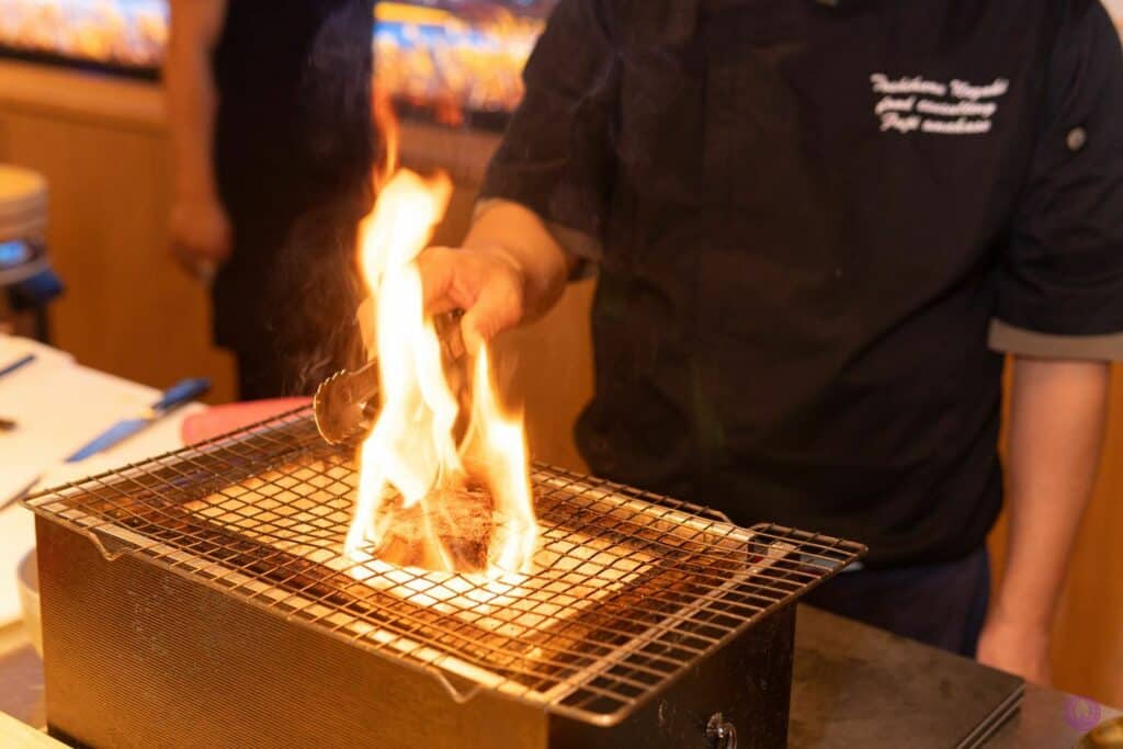 A chef in a dark uniform uses tongs to cook Japanese Wagyu over an open flame on a wire grill placed atop a rectangular metal stove. Bright flames rise from the grill, and the chef&rsquo;s face is not visible in the frame. Fuji Omakase Sushi San Fernando Valley