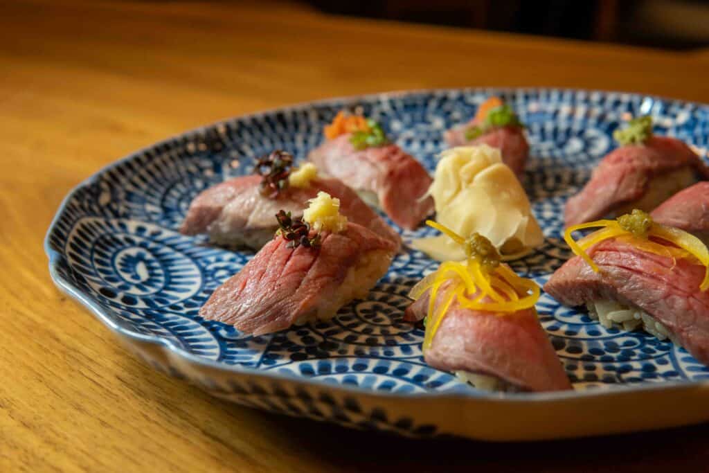 A decorative blue-and-white plate holds several pieces of nigiri sushi topped with slices of marbled Japanese Wagyu beef, each garnished with various toppings. Pickled ginger is placed in the center, and the background is softly lit wood. Fuji Omakase Sushi San Fernando Valley